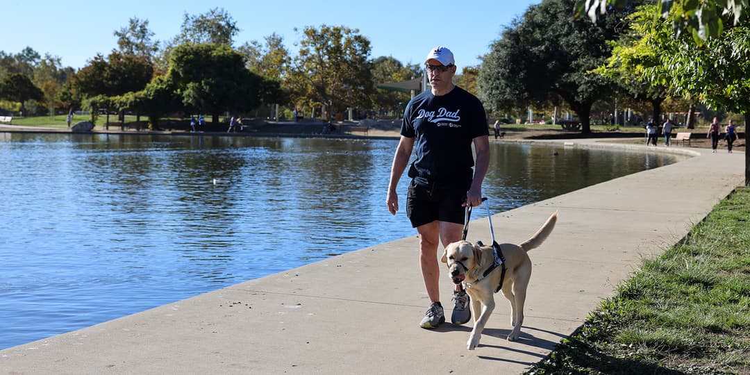Man in shorts walking with dog on sidewalk along water